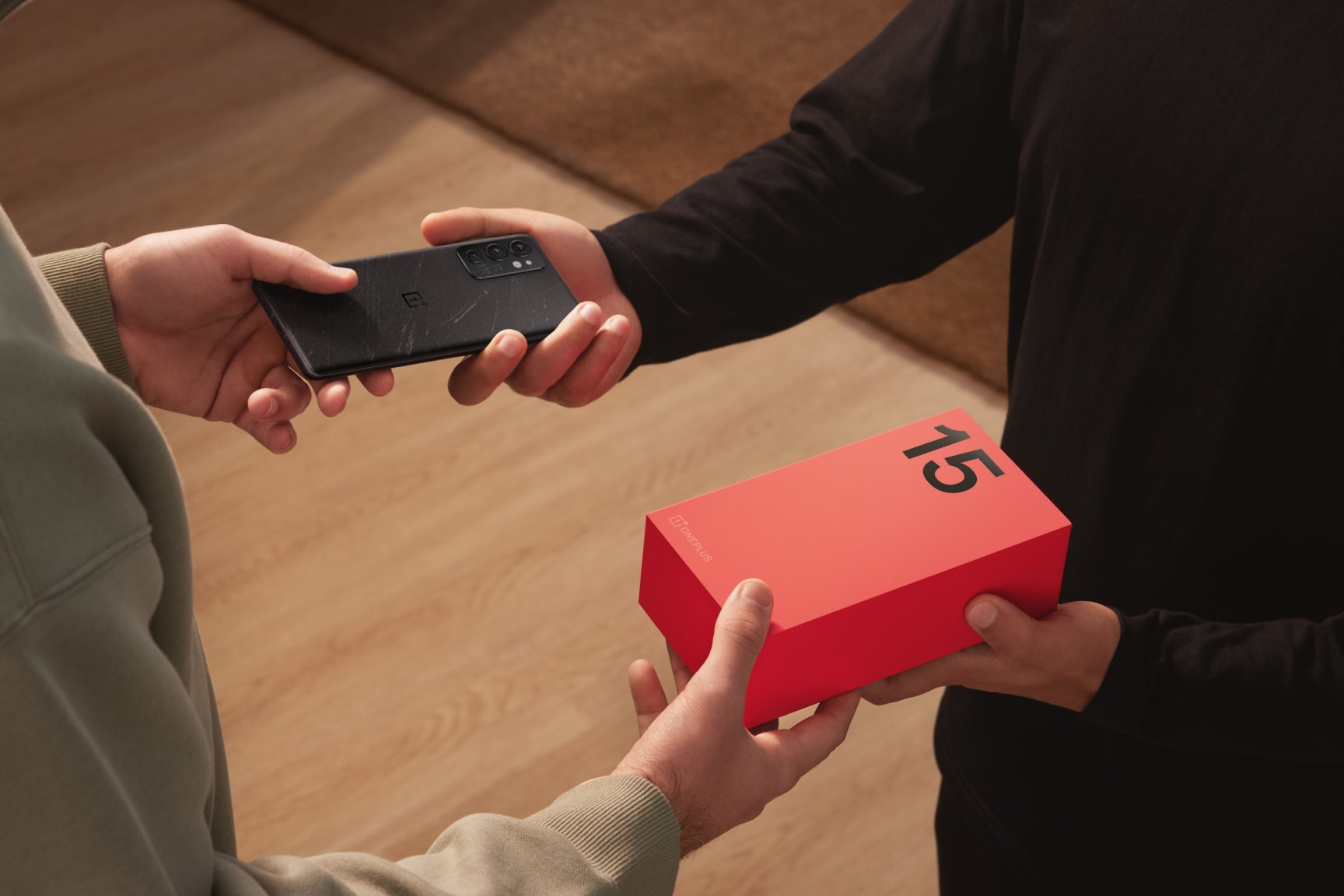 Two people exchanging a black mobile device and a red box on a wooden floor.