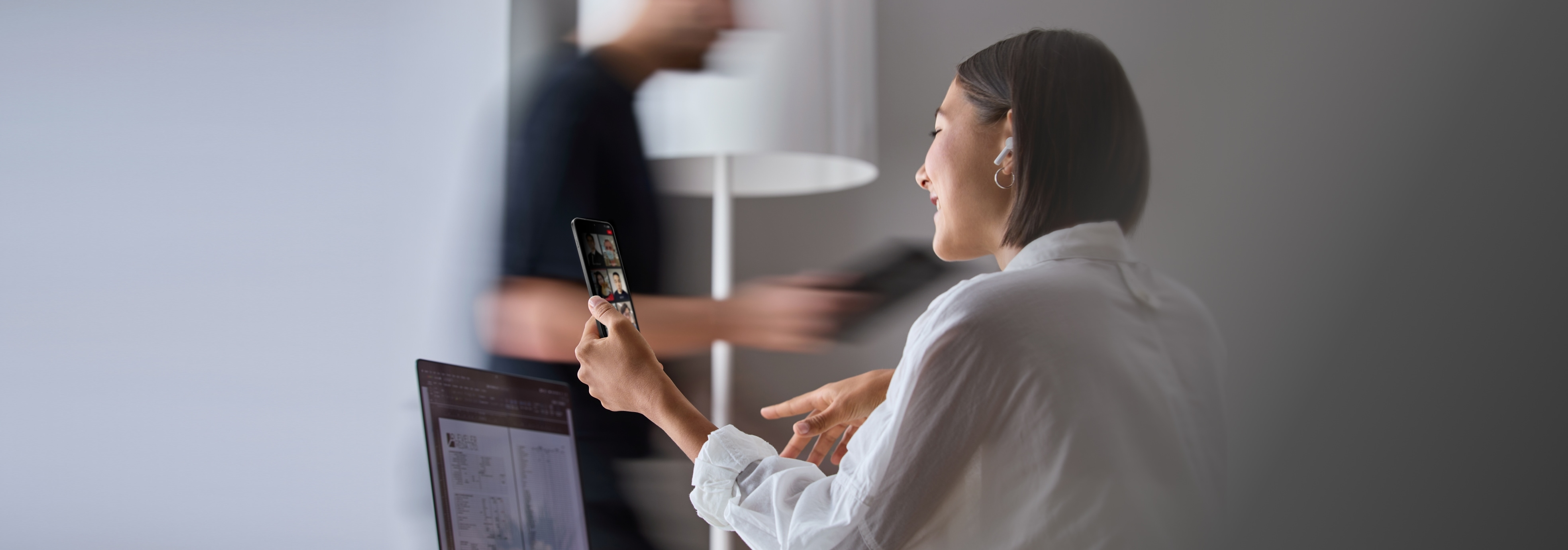 A woman in a white shirt using a smartphone while working on a computer in a modern office.