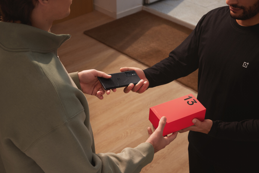 Two people exchanging a black mobile device and a red box on a wooden floor.